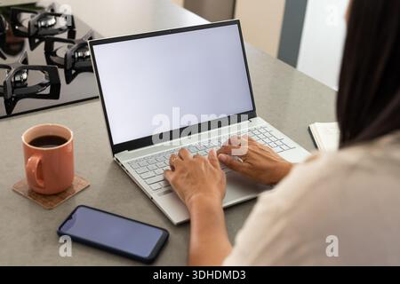 Laptop resting on kitchen counter with blank screen, coffee mug, smartphone, notebook, stovetop Stock Photo