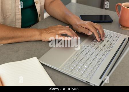 Hands are typing on silver laptop with backlit keys on gray countertop, coral mug nearby Stock Photo