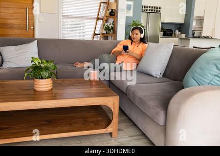 Senior woman lying on sofa in living room wearing orange shirt headphones holding phone, copy space Stock Photo