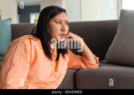 Mature woman sitting floor at home leaning against gray sofa wearing peach shirt gazing at window Stock Photo