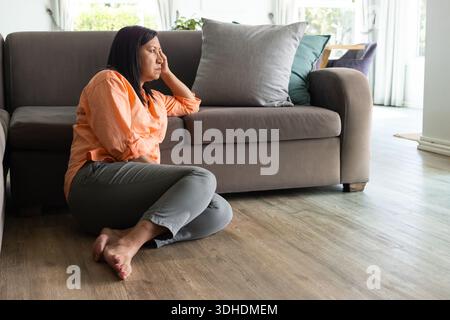 Mature adult woman sitting on floor leaning on gray sofa wearing orange blouse gray pants barefoot Stock Photo