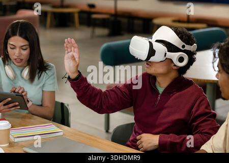 African American students sitting in campus lounge around table using VR headset, tablet, laptop Stock Photo