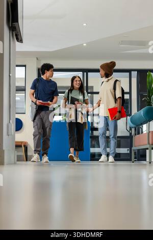 Diverse students walking and talking down campus hall holding backpacks notebooks bright red folder Stock Photo