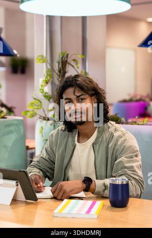 Indian man wearing green bomber jacket sitting at wooden table in cafe, using tablet and pen Stock Photo