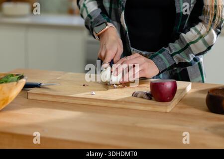 Garlic bulb is being sliced on wooden cutting board at kitchen counter, red onion nearby Stock Photo