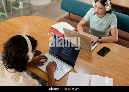 African American peers wearing white headphones, woman in mint-green top, typing on laptops in cafe Stock Photo