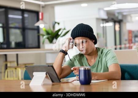 African American man in black beanie resting head, focusing on tablet at cafe table with notebook Stock Photo