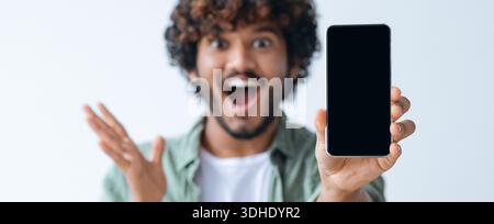 Excited Young Man Shows Smartphone in Bright Setting While Smiling and Gesturing Stock Photo