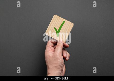 Hand holding a wooden card with a green checkmark icon on a neutral gray background. Business approval, confirmation, quality control and successful d Stock Photo