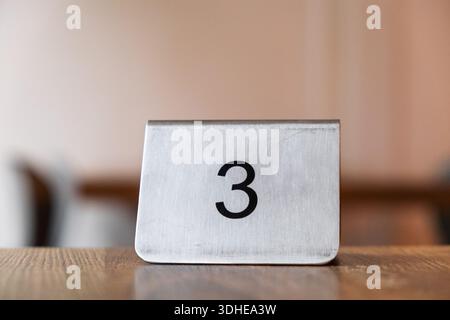 A metal table number '3' sits on a wooden surface, likely in a restaurant or cafe, indicating a table assignment for patrons. Stock Photo