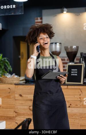African American woman in black apron laughing and talking on phone holding tablet at cafe counter Stock Photo
