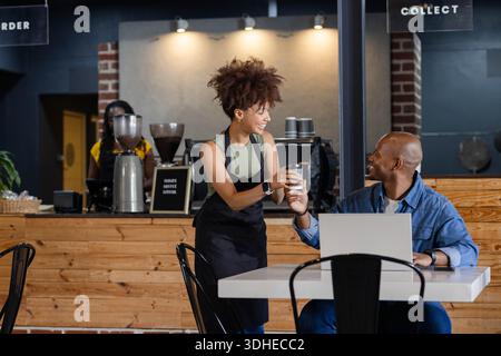 Female barista wearing black apron handing takeaway coffee cup to seated man with laptop at counter Stock Photo