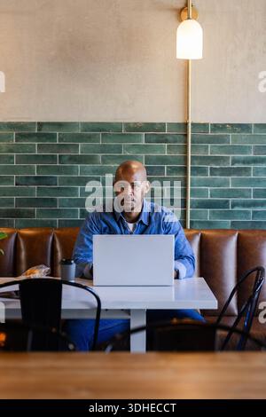 African American man sitting in denim shirt at cafe working on silver laptop with coffee cup Stock Photo