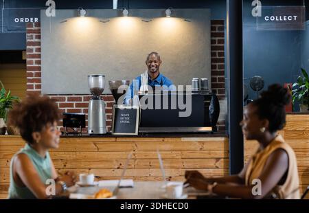 African American barista in apron at cafe counter pulling espresso while women typing on laptops Stock Photo