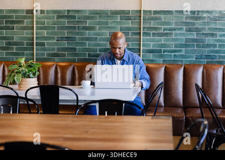 African American adult man sitting in quiet cafe booth, working on silver laptop with coffee cup Stock Photo