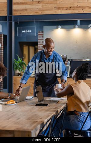 African American male server in denim-shirt black-apron handing cup at communal table with laptops Stock Photo