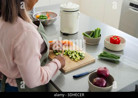 Zucchini being sliced on wooden cutting board on gray countertop, with knife and compost canister Stock Photo