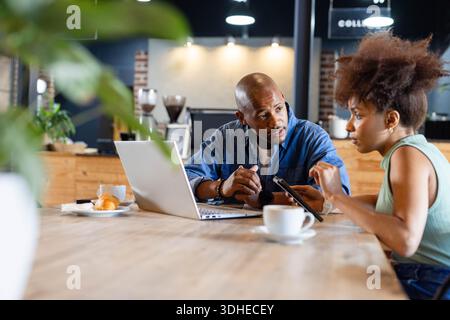Colleagues sitting at coffee shop table, discussing tablet while using laptop, cups, croissant Stock Photo