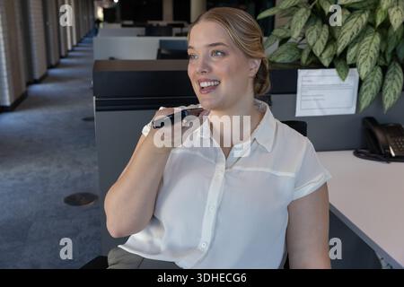 Female office worker in white blouse holding phone speaking at desk with desk phone and plant Stock Photo