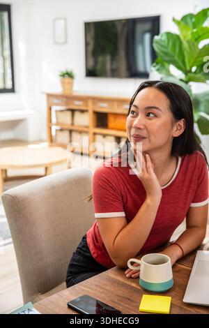 Asian woman sitting at home table wearing redtop thinking near mug phone laptop notes, copy space Stock Photo