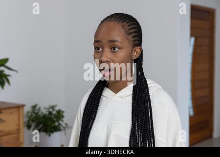 African American woman standing against light wall at home wearing hoodie and touching long braids Stock Photo