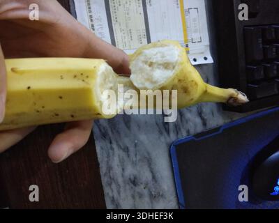 Hand holding a partially peeled banana on a desk with keyboard and mouse Stock Photo