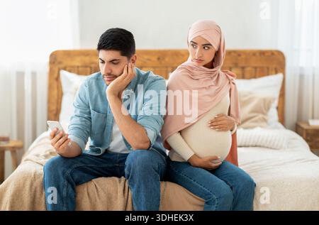 Young pregnant muslim woman offended to her husband busy with smartphone, arab lady in hijab feeling lonely Stock Photo
