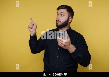 Man in black shirt holding smartphone, pointing upward against yellow background. Stock Photo