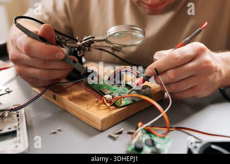 Close-up view of technician focusing on intricate details of circuit board on wooden base with magnifying glass, using helping hands tools and wires Stock Photo
