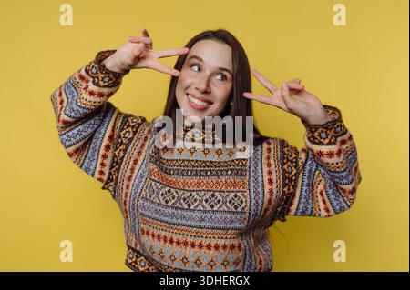 Smiling woman in patterned sweater making peace signs against yellow background. Stock Photo
