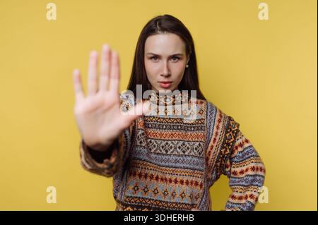 Woman in patterned sweater making stop gesture on yellow background Stock Photo