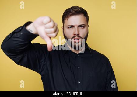 Man in black shirt giving thumbs down against yellow background. Stock Photo