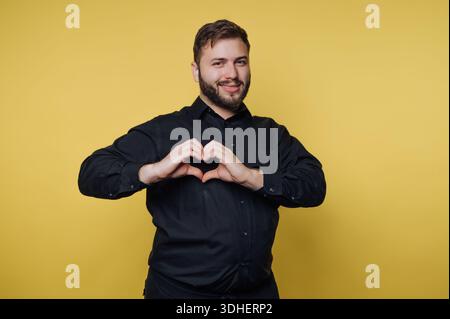 Man in black shirt making heart shape with hands on yellow background. Stock Photo