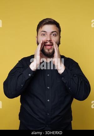 Man in black shirt shouting with hands near mouth on yellow background. Stock Photo
