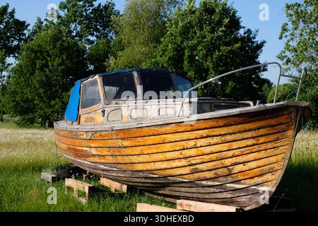 An old, weathered wooden motorboat, named Elvira, is dry-docked on wooden blocks on a grassy lawn Stock Photo