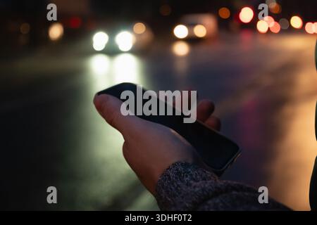 Close up of hand holding smartphone at night with blurred city traffic lights in background creating modern urban lifestyle and technology concept. Stock Photo