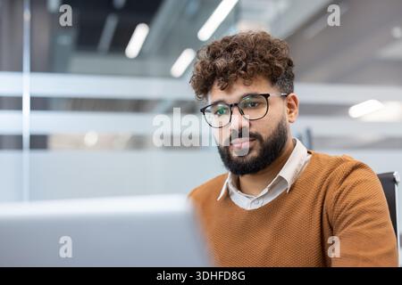 Young professional man with curly hair, beard and glasses intently working on a laptop in a modern office, focused on strategy, productivity and digital development Stock Photo