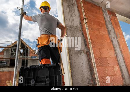 A worker stands on a crate, looking at building walls. Tools and equipment are visible on the construction site. Stock Photo