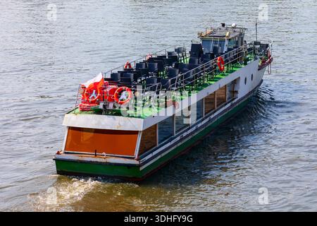 Passenger boat with superstructure navigates on the water, open upper deck with many dark chairs and visible flag. Vessel is pushing wake as it moves, Stock Photo