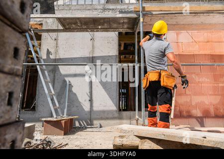 A construction worker stands on a site working on wall materials while wearing a yellow hard hat and safety gear. Stock Photo