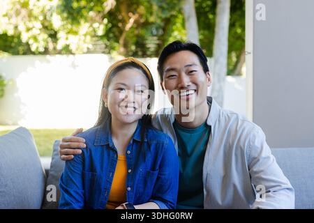 Asian couple sitting on light-gray sofa in covered patio, smiling at camera with yellow headband Stock Photo