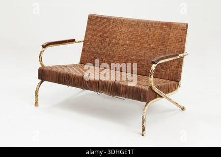A vintage woven rattan bench with a metal frame and curved armrests, showing signs of age and patina, isolated on a light grey background Stock Photo