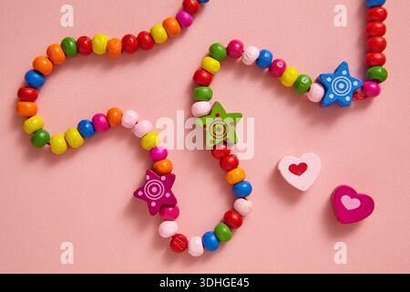 A colourful handmade beaded necklace featuring bright rainbow beads with cute star and heart charms, arranged on a soft pink background. Stock Photo