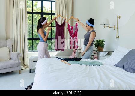Two women are comparing dresses on wooden hangers in bedroom, window light filling room Stock Photo