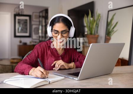Happy young Indian woman in wireless headphones writing notes Stock Photo