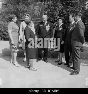 Slavyansk, Donetsk region, USSR. 1980s. A lively conversation between World War II veterans and factory workers at the AIZ plant memorial. An elderly veteran with many medals smiles broadly while talking to a group of women standing on the left. On the right, other colleagues listen to the story with interest. Behind them is the granite monument with a soldier's portrait. This archival black and white photo captures the atmosphere of friendship, joy of life, and generational connection during the Victory Day celebration in the peaceful Donbass. Stock Photo