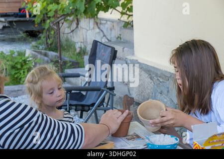 Two children work with clay pots, shells, and paint at a family DIY station under a terrace. Stock Photo