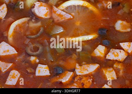 Classic solyanka bowl with lemon and brined olives, smoky meat cubes, diced potato immersed in rich spiced tomato broth, rustic presentation suggestin Stock Photo