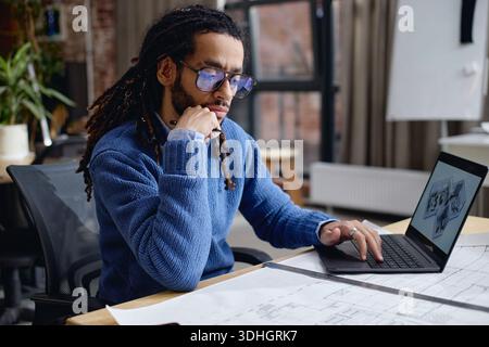 Young Adult Black Man Working on Architectural Plans Using Laptop Computer Stock Photo