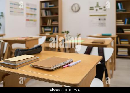 Empty classroom showing wooden desks with notebooks, pens, and pencil case arranged neatly, bookshelves filled with books in background, educational posters and clock on wall Stock Photo